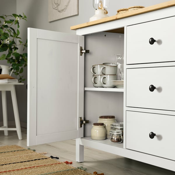 Buffet blanc HEMNES avec porte ouverte révélant les tasses, bocaux et assiettes rangés. Expose soigneusement les objets ménagers.