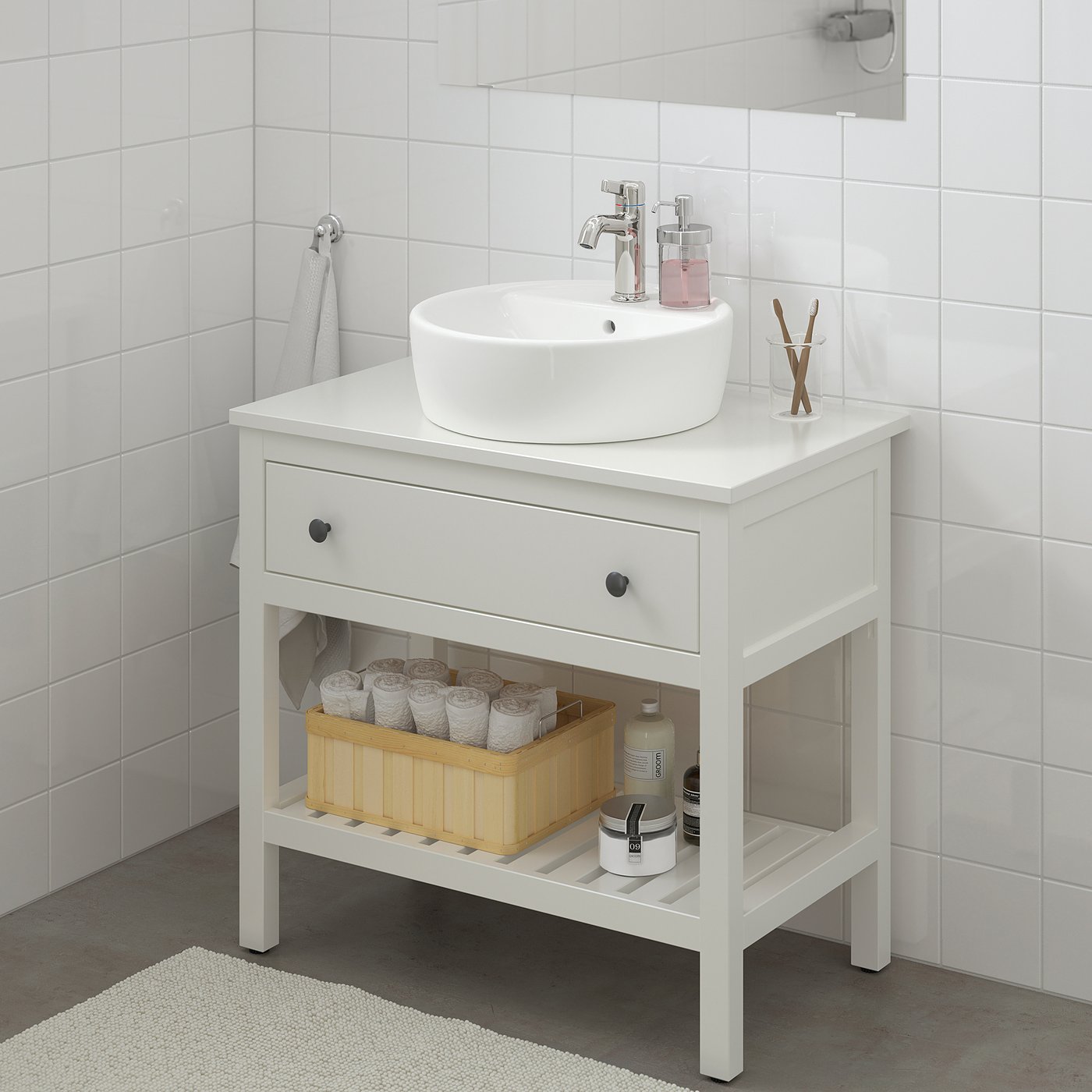 Vanité HEMNES blanche avec évier rond, tiroir et étagère. Le panier contient du papier toilette et du shampoing, une serviette grise à proximité.