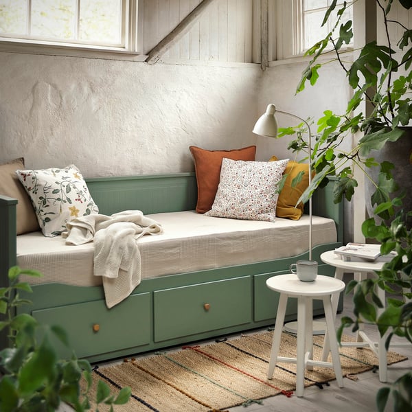 Lit de jour HEMNES en gris-vert avec literie crème, mug et lampe sur la table d’appoint, tapis rayé. Moderne et confortable.