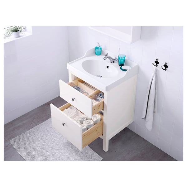 Vanité de salle de bain blanche avec tiroirs ouverts révélant des serviettes et des articles de toilette soigneusement empilés. Deux poignées visibles sur le mur.