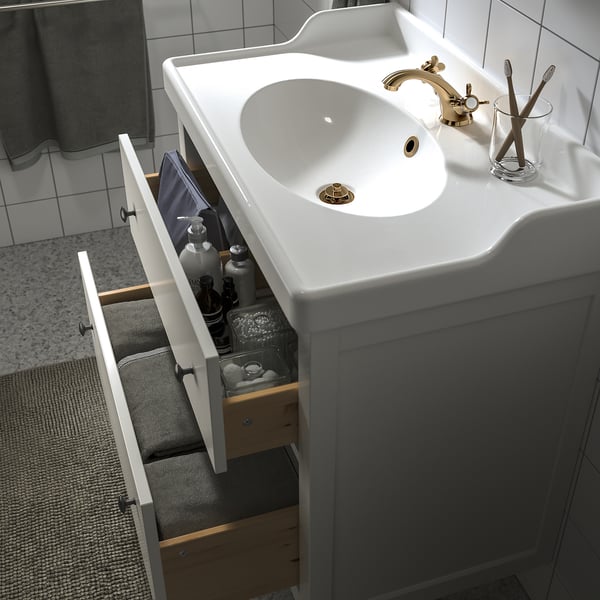 Meuble de salle de bain blanc avec tiroir ouvert montrant les articles de toilette et les serviettes, robinet doré, porte-brosse à dents.