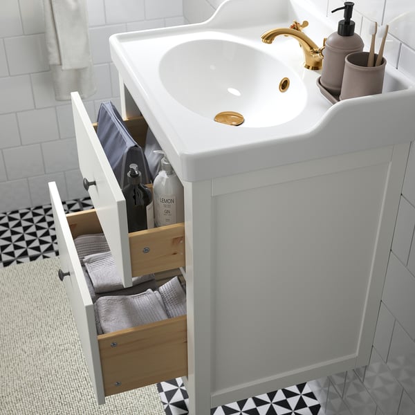 Lavabo de salle de bain moderne avec tiroir ouvert montrant des serviettes et des articles de soins capillaires soigneusement pliés. Luminaires dorés et carrelage géométrique au sol visibles.