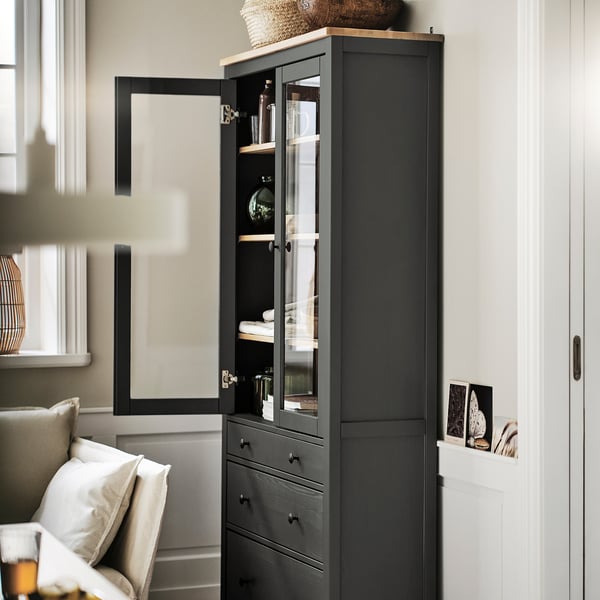 Un placard HEMNES sombre avec des portes vitrées, dressé haut à côté d’une porte. Elle est en bois massif, haute avec des étagères réglables, rangeant soigneusement la verrerie.