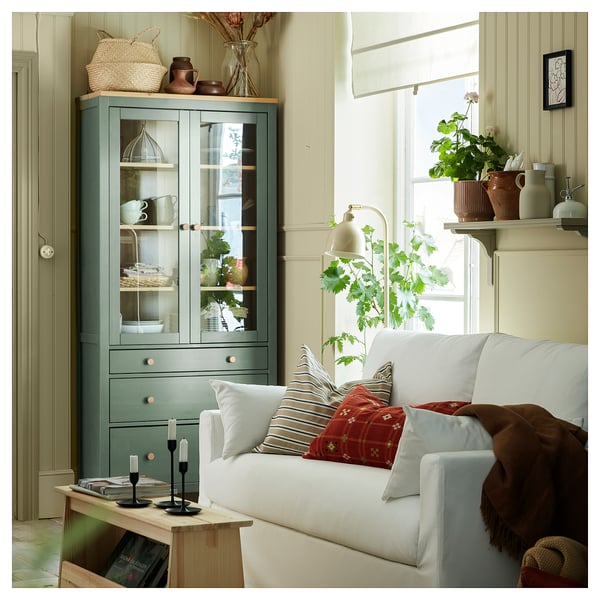 Salon avec canapé blanc, oreillers colorés, armoire en bois vert, étagères avec plantes et décorations, fenêtre avec rideau léger.