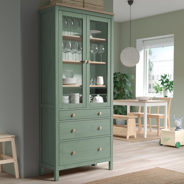 Armoire verte HEMNES avec portes vitrées, rangeant la vaisselle et les verres dans une salle à manger en bois clair.