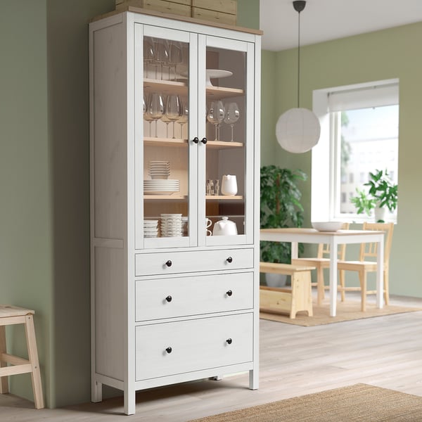 Armoire en bois HEMNES avec portes vitrées et banc contre un mur vert, rangeant la vaisselle. Contexte : table à manger, chaises et fenêtre.
