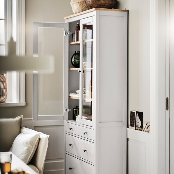 Un grand meuble en bois HEMNES, blanc, avec des portes et des tiroirs en verre. Elle expose des objets décoratifs à l’intérieur et se trouve près d’un escalier et d’une chaise.