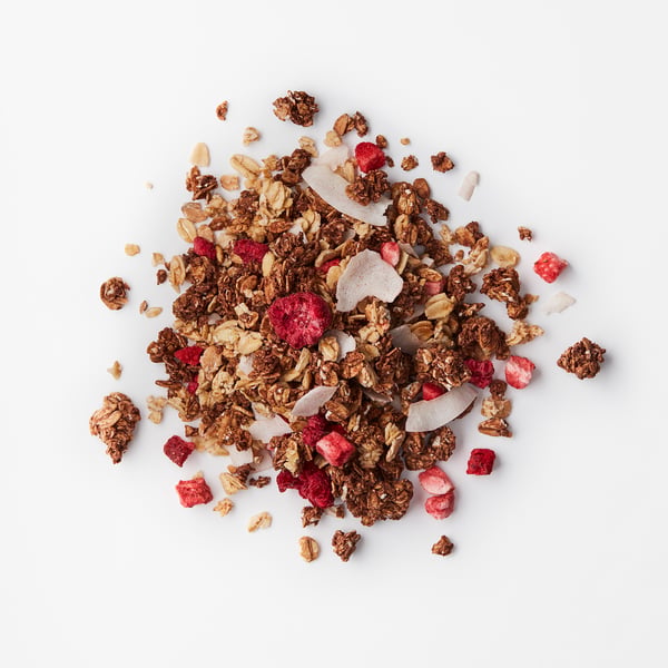 Mélange de granola bombé avec des flocons d’avoine au chocolat croquants, des flocons de noix de coco et des baies séchées rouge vif sur une surface blanche.