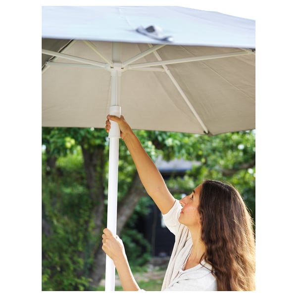 Une femme ajuste un parasol HÖGÖN beige à l’extérieur, en veillant à ce que l’ombre se produise.