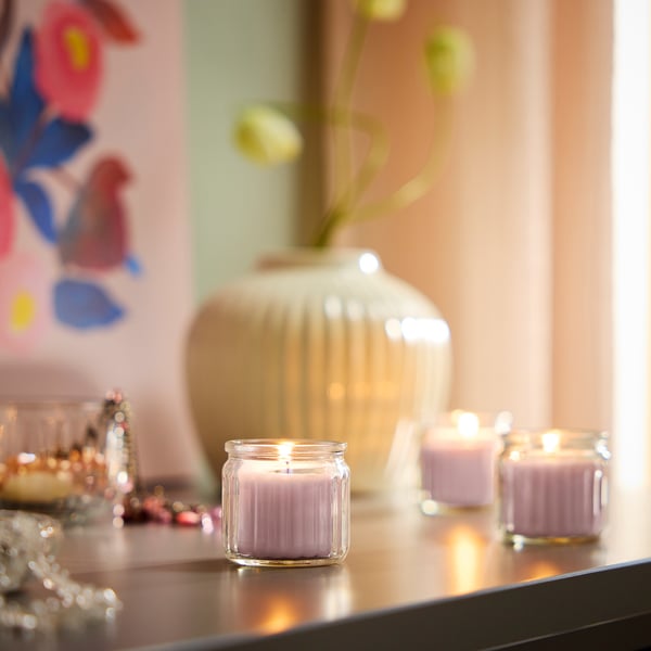 De petites bougies roses dans des boîtes en verre sur une table, entourées d’objets décoratifs comme un vase avec des fleurs et un oiseau peint.