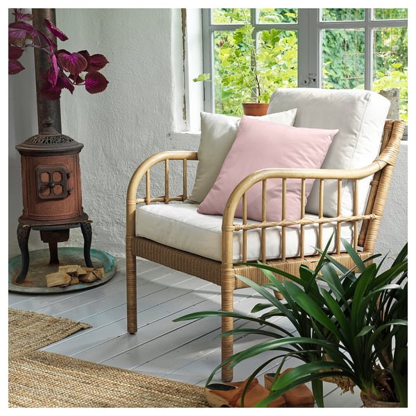 Un fauteuil en osier HOLMSTA avec des coussins beiges et roses. Il utilise des matériaux naturels tels que le rotin et le bambou, créant un design confortable et rustique.