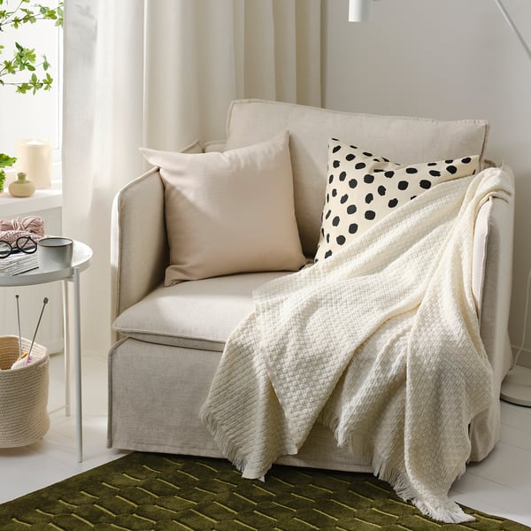Plaid beige HORNMAL sur fauteuil, oreillers, table d’appoint avec tasse, tricot, plante, tapis vert - cadre confortable.
