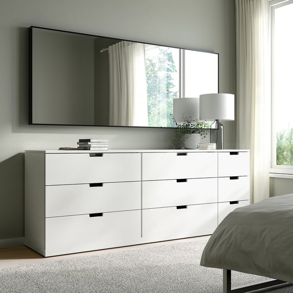 Miroir HOVET blanc au-dessus de la commode large dans la chambre lumineuse. Le miroir reflète la verdure, la commode contient des livres, une lampe et une plante.