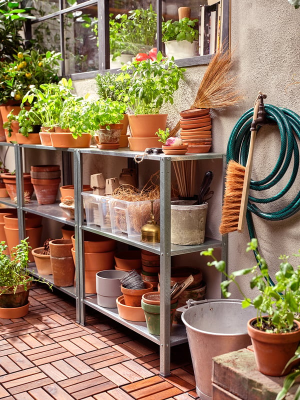 Stockage extérieur des plantes : étagère HYLLIS en acier galvanisé contenant des pots en terre cuite, des outils de jardinage et des plantes. Tuyau vert et brosse attachés.
