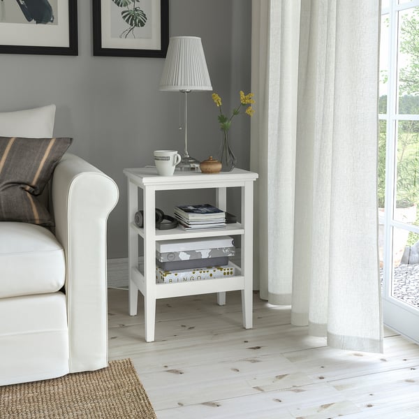Petite table d’appoint blanche IDANÄS à côté du canapé, contenant une lampe, des tasses et des livres. Parquet clair, mur gris, rideaux transparents.