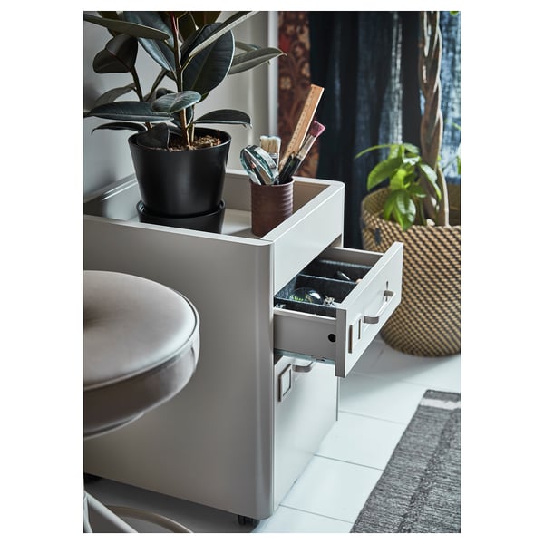 Un meuble à tiroirs IDÅSEN gris avec une plante en pot et des articles de toilette sur le dessus, situé dans une salle de bains avec des rideaux sombres.