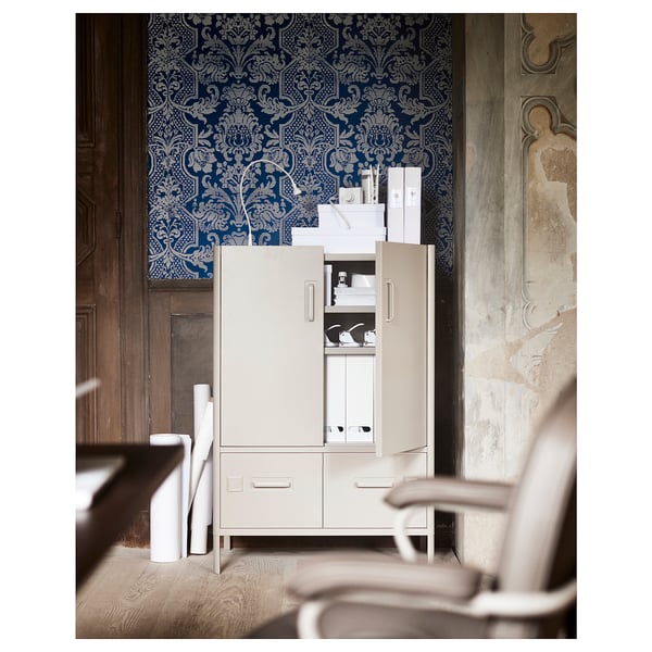 Une armoire de bureau IDÅSEN blanche avec un fond de papier peint à motifs bleus. L’armoire a des tiroirs ouverts et des portes révélant des objets de rangement.