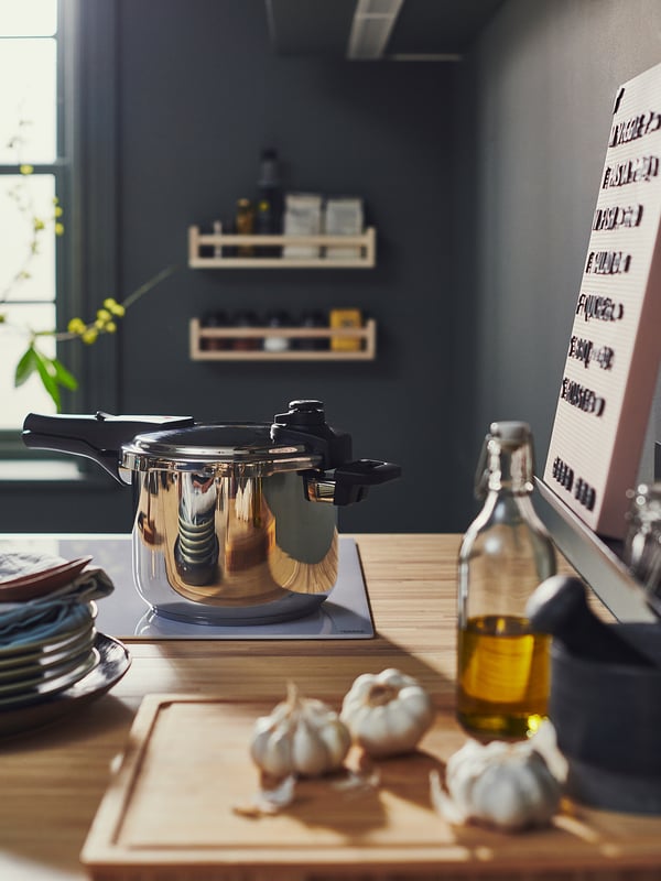 Comptoir de cuisine moderne avec autocuiseur en acier inoxydable IKEA 365+, ail, bouteille d’huile et vaisselle, mur sombre avec étagères.