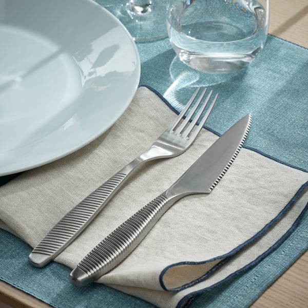 Mise en place de la table avec couverts IKEA 365+ : fourchette et couteau dentelé sur une serviette de table, à côté d’une assiette blanche et d’un verre.