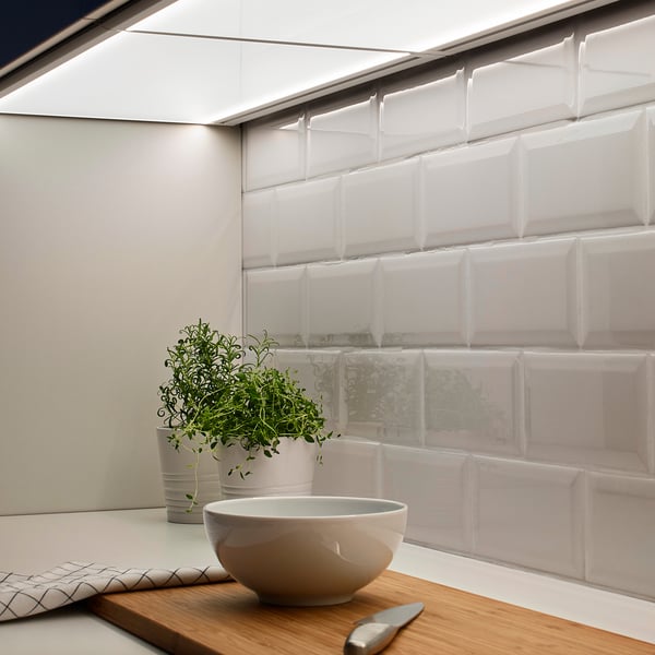 Comptoir de cuisine moderne avec bol blanc, couteau et herbes en pot sur une planche de bois près du panneau lumineux sous l’armoire.