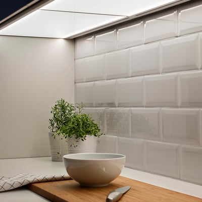 Comptoir de cuisine moderne avec bol blanc, couteau et herbes en pot sur une planche de bois près du panneau lumineux sous l’armoire.