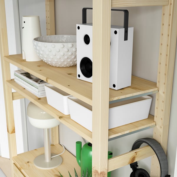 Unité de rangement en bois avec étagères contenant un haut-parleur blanc, un bol blanc, des récipients blancs, des livres, un arrosoir vert et une lampe blanche.