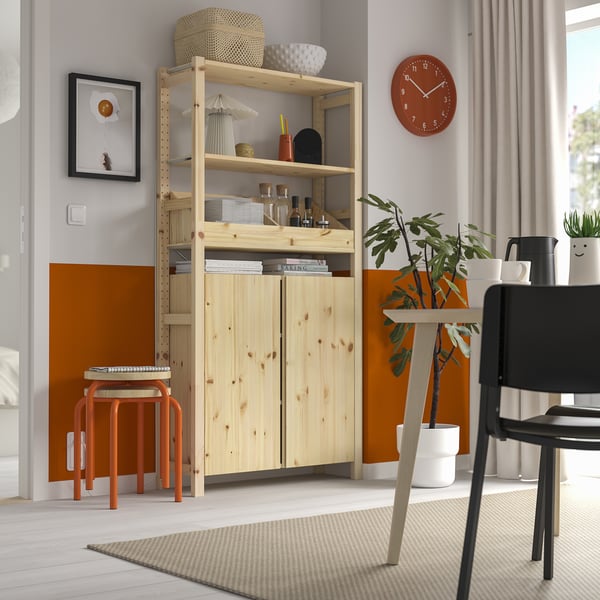 Bibliothèque IVAR en pin avec étagères et armoires. Horloge rouge, plante et chaise. Tabouret orange, tapis tissé.