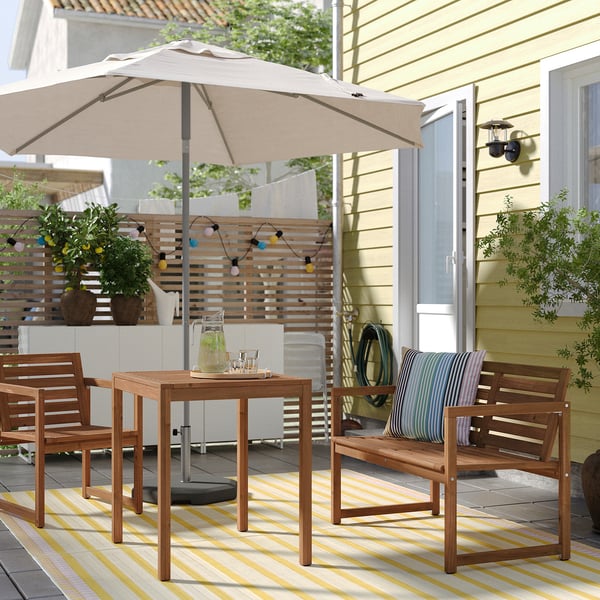 Scène de patio confortable avec table en bois, chaises, parasol beige, tapis rayé, lumières colorées et plantes en pot.
