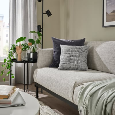 Salon avec coussin JORDKASTANJ gris, ordinateur portable, livres, plante et décor minimaliste.