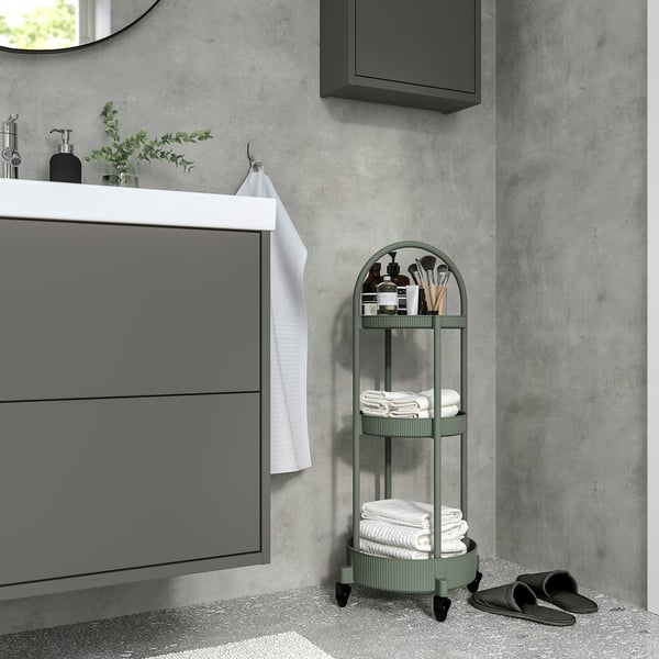 Un chariot roulant gris-vert à trois niveaux nommé JUTTERSBO à côté d’un lavabo de salle de bain. Il peut contenir des serviettes et des articles de toilette, s’adaptant bien aux petits espaces.