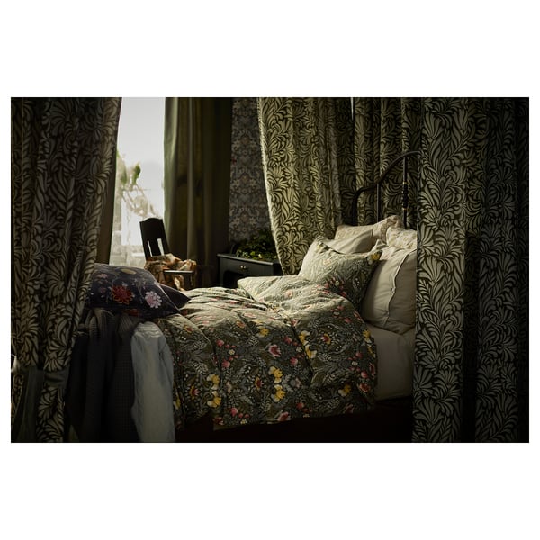 Lit avec couette florale, oreillers crème, rideaux sombres, table de chevet et plante.