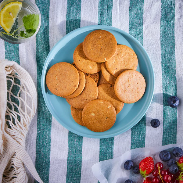 Une assiette bleue de biscuits au gingembre brun repose sur un tissu rayé vert, à côté d’une boisson au citron et à la menthe, et d’un panier de baies mélangées.