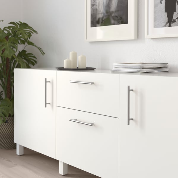 Armoire blanche avec poignées métalliques, deux tiroirs et deux portes. Décoré de bougies et de livres, à côté d’une plante.