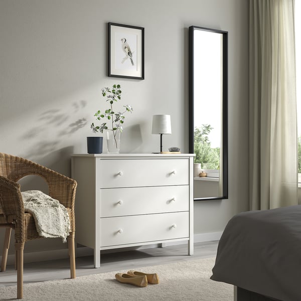 Chambre moderne avec coffre KILBERGET blanc, miroir, lit gris, chaise en osier, petite lampe, plante et art mural blanc.