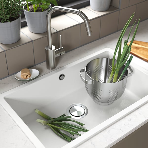 Évier de cuisine avec passoire en acier inoxydable, oignons verts, robinet et plantes en pot.