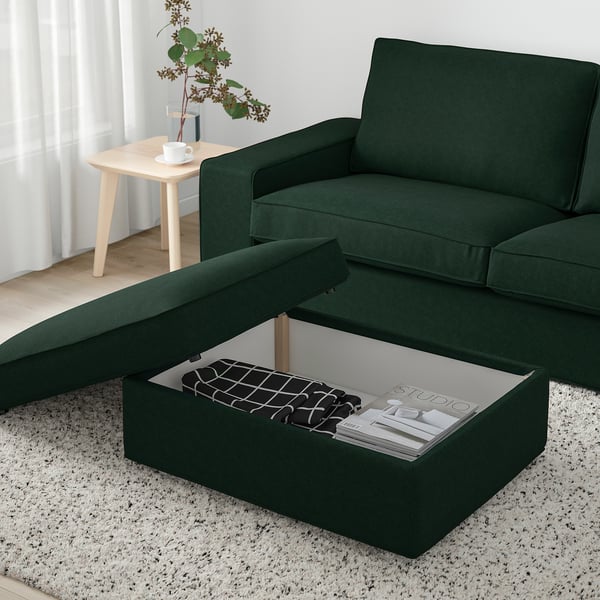 Repose-pieds KIVIK vert avec rangement ouvert, montrant des magazines, sur tapis blanc près de la fenêtre.
