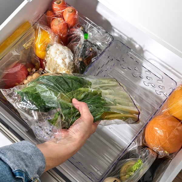 La main d’une personne tenant un paquet de légumes emballé dans un plastique dans un tiroir du réfrigérateur. Le tiroir est organisé avec divers fruits et légumes dans un plastique transparent.