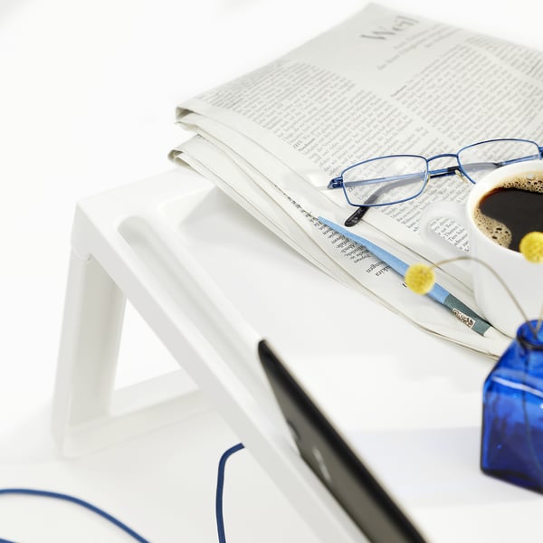 Un plateau de lit pliable blanc contient un journal, des lunettes, du café et un stylo, illustré par une tablette partiellement visible et un fond blanc.