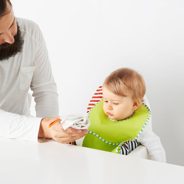 Homme nourrissant bébé avec un bavoir vert à l’aide d’un gant de toilette.