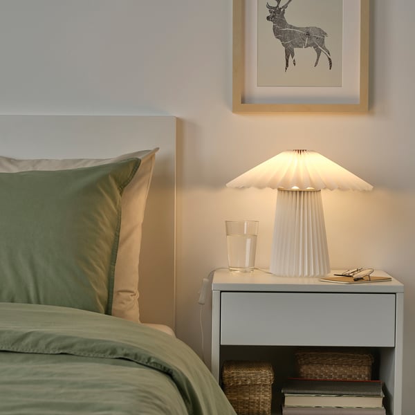 Une chambre confortable avec une lampe KUDDLAVA sur une petite table, avec un oreiller vert, des livres et une photo de cerf.