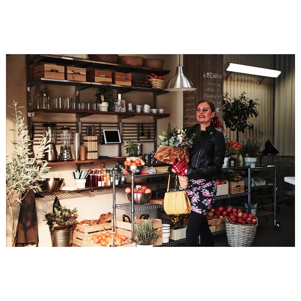 Profitant de l’épicerie, une femme en robe fleurie et veste en cuir se tient dans une cuisine rustique avec des caisses en bois, des produits frais et des paniers suspendus.