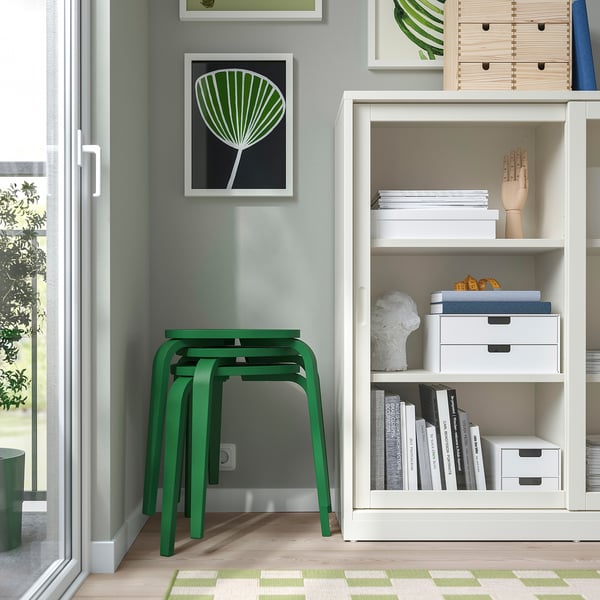Chambre moderne avec des tabourets verts, une bibliothèque blanche remplie de livres et d’objets décoratifs, et une impression d’art abstrait encadrée sur le mur.