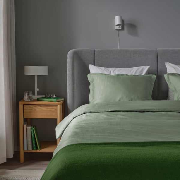 Lit gris, couette verte, oreillers blancs. Table de chevet en bois, lampe, livres. Applique. Chambre confortable.