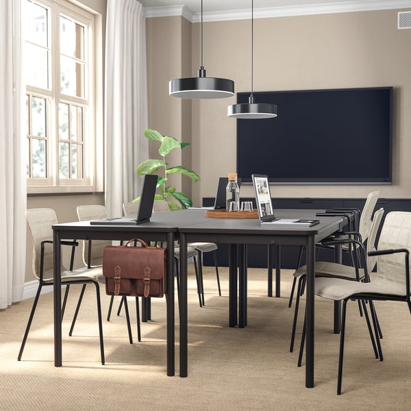 Salle de conférence moderne avec tables sombres, chaises, télévision et lampes suspendues. Lumière naturelle et décor végétal.