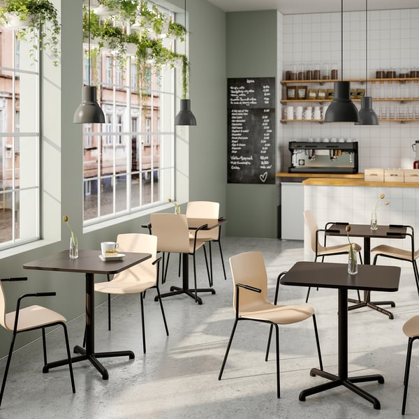Café moderne avec tables en bois et chaises beiges. Des plantes suspendues, de grandes fenêtres, des lampes noires et un menu à l’ardoise créent une atmosphère détendue.
