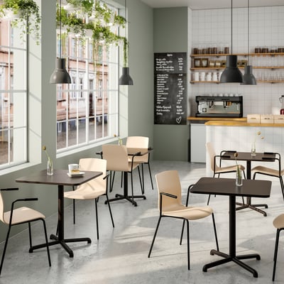 Café moderne avec tables en bois et chaises beiges. Des plantes suspendues, de grandes fenêtres, des lampes noires et un menu à l’ardoise créent une atmosphère détendue.