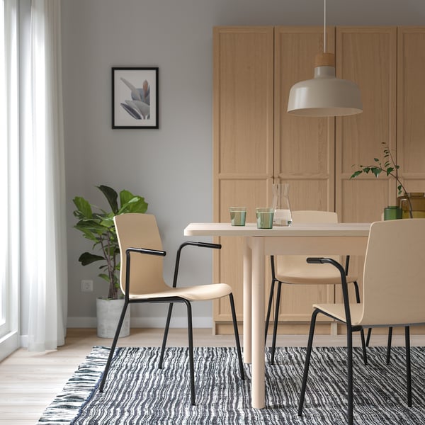 Salle à manger moderne avec table en bois clair, chaises LÄKTARE en bouleau, pieds en métal noir, tapis et suspension. Murs gris, plantes et art.