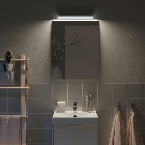 Petit miroir LÄRBRO gris dans la salle de bains, éclairé au-dessus d’un lavabo blanc, entouré de carreaux sombres.