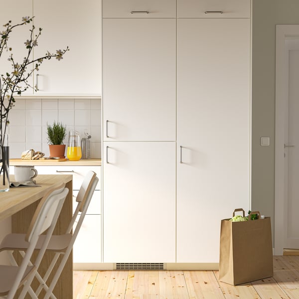 Un congélateur de cuisine intégré compact blanc, légèrement plus petit que de grandes armoires, placé à côté de chaises et d’un sac d’épicerie.