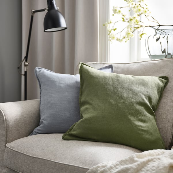 Salon confortable avec coussin gris-vert sur canapé beige, oreiller bleu, lampe et fleurs blanches.